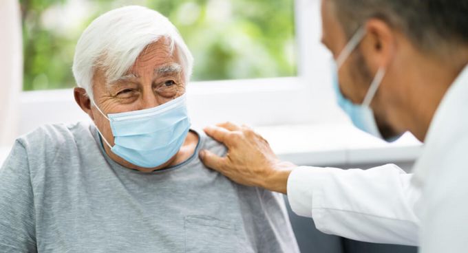 Arzt mit Maske legt einem älteren Patienten mit Maske die Hand auf die Schulter